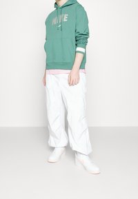 Grön bomulls-HOODIE med framficka, grå logotyp, över en rosa tröja. Vita cargobyxor och vita sneakers med rosa detaljer.