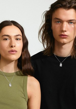Collane d'argento con pendenti rettangolari indossate da due persone; una con una maglietta verde oliva, l'altra con una camicia nera. Texture lisce.