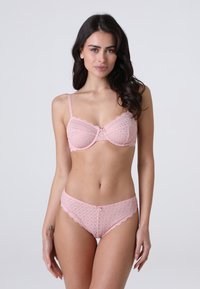 Donna con lunghi capelli scuri che indossa un reggiseno di pizzo rosa e mutandine abbinate, in piedi contro uno sfondo chiaro e uniforme, rivolta in avanti.