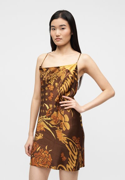 Νεαρή γυναίκα με μακριά σκούρα μαλλιά φορώντας καφέ και χρυσό φόρεμα με λουλουδένιο σχέδιο και λεπτές τιράντες, ποζάροντας με το ένα χέρι στη μέση.