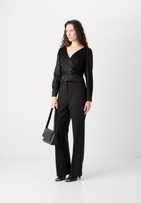 Top avvolgente nero a maniche lunghe con texture floreale, abbinato a pantaloni neri a vita alta e scarpe nere con la punta. Porta una borsa a tracolla nera.