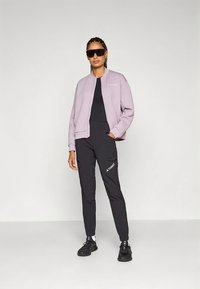 Lys lilla quiltet bomberjakke med svarte bukser med logo. Sorte sko og solbriller fullfører antrekket. Enkel, funksjonell design.