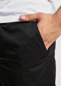 Pantalons noirs avec une texture lisse et mate, dotés d'une poche latérale et d'une coupe ajustée, complétés par un t-shirt blanc à la taille.