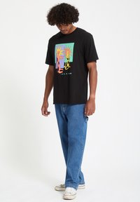 Svart bomull T-skjorte med et fargerikt grafisk print med sol- og figurdesser. Kombinert med blå denim bukser og hvite joggesko.