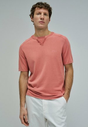Jeune homme aux cheveux bouclés portant une chemise henley corail à manches courtes et un pantalon blanc, debout avec une main dans la poche.