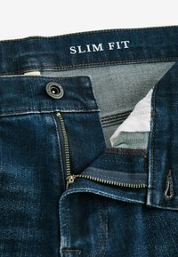 Donkerblauwe slim fit jeans met een ritssluiting en knoopsluiting. Het interieur is voorzien van beige stof en contrasterende stiksels.