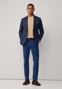 Blazer a scacchi blu navy sopra un maglione beige, abbinato a pantaloni blu e scarpe stringate marroni. Caratteristiche includono due tasche frontali e un fazzoletto da taschino.