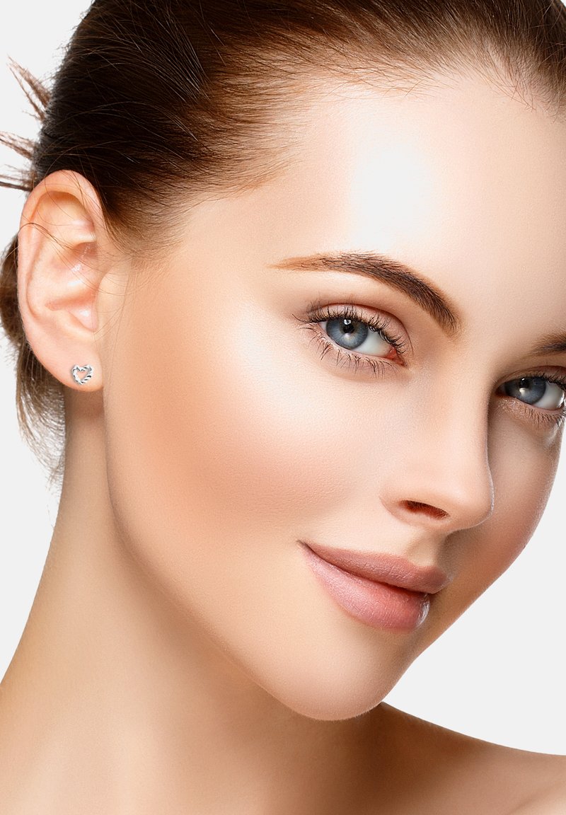 Närbild av ett ansikte med slät hy, prydd med lätt smink. Silverfärgade hjärtformade studs pryder örat. Ljusbrunt hår.