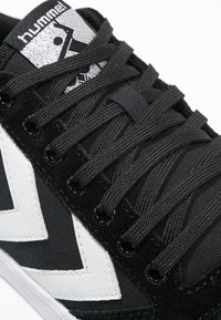 Musta sneaker, jossa on valkoinen viistoviiva-kuvio, mustat nauhat, jotka on pujotettu metallisiin reikäkohdiin, sekä hopeinen logopatch kielen päällä.