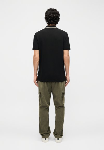 Άνδρας με σγουρά μαλλιά στέκεται γυρισμένος, φοράει μαύρο κοντομάνικο πόλο πουκάμισο, ελαιοπράσινο παντελόνι cargo και λευκά αθλητικά παπούτσια.