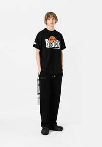Camiseta de algodón negra con texto y gráfico en blanco, combinada con pantalones de chándal negros con un bolsillo lateral y zapatos negros de textura tipo slip-on.