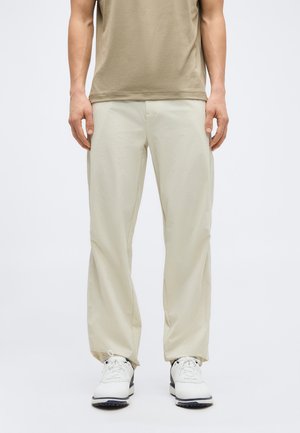 Homme portant un pantalon beige décontracté, une chemise à manches courtes couleur bronze et des baskets blanches sur un fond clair uni.