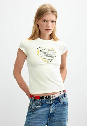 Femme blonde portant un t-shirt blanc cintré avec un texte imprimé en forme de cœur et un jean bleu avec une ceinture à motifs, debout sur un fond blanc.