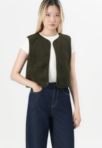 Olivenfarget fleecevest med trykknapper, rund halslinje, ermeløs design, brukt over en hvit T-skjorte og paret med mørkeblå jeans.