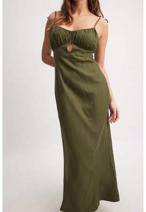 Mujer con un vestido largo verde oliva con tirantes finos y un pequeño corte en forma de ojo de cerradura debajo del busto, posando sobre un fondo liso.