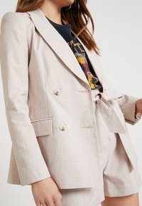 Femme portant un blazer beige à carreaux à double boutonnage et un short taille haute assorti noué, sur un t-shirt noir à motif.