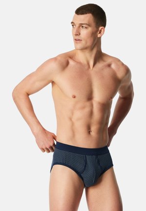 Skjorteløs mand justerer marineblå mønstrede underbukser, kigger mod venstre, foran en ensfarvet lysegrå baggrund.