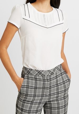 Femme portant un chemisier blanc à manches courtes avec bordure noire et plis, associé à un pantalon à carreaux noir et blanc taille haute.