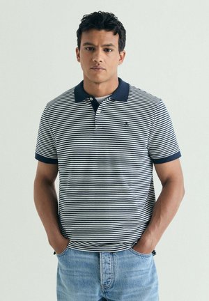 Joven con cabello corto y rizado, vestido con un polo de rayas azul marino y blanco y vaqueros azul claro con las manos en los bolsillos.