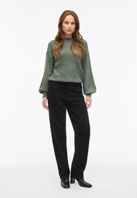 Zielony sweter o fakturowanej powierzchni z długimi rękawami, połączony z czarnymi sztruksowymi spodniami i czarnymi botkami, stojący na białym tle.