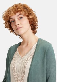 Cardigan verde su una maglietta beige con ricami intricati, con scollatura a V. La texture morbida e il tessuto leggero sono evidenti.