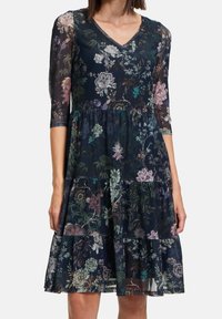 Robe imprimée florale en marine avec des manches transparentes trois-quarts, un décolleté en V et une jupe à volants. Présente un mélange de couleurs pastel sur le tissu.