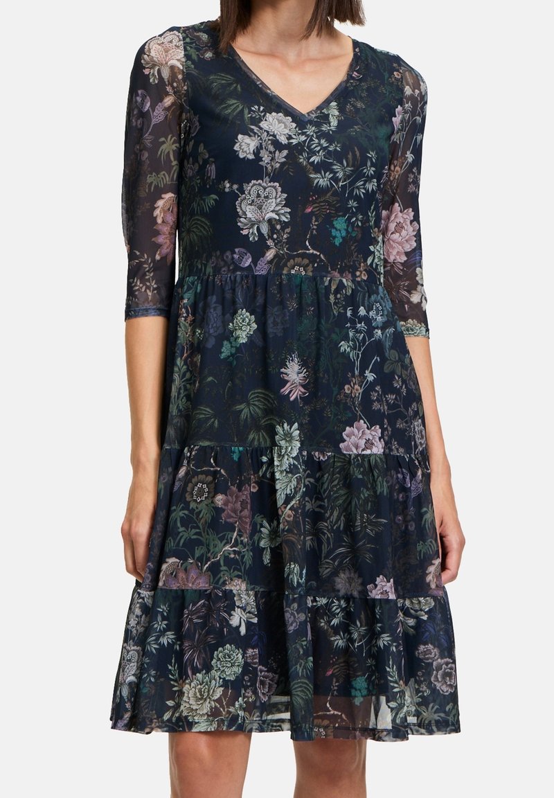 Robe imprimée florale en marine avec des manches transparentes trois-quarts, un décolleté en V et une jupe à volants. Présente un mélange de couleurs pastel sur le tissu.
