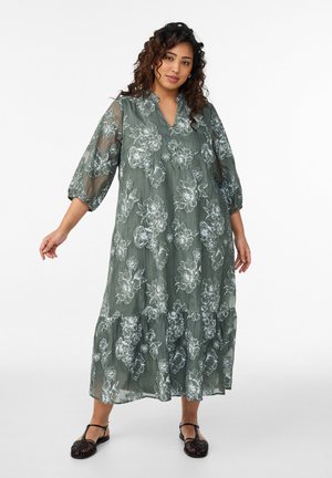 Mujer con un vestido largo de gasa verde con patrones florales blancos y mangas abullonadas hasta el codo, de pie con los brazos ligeramente extendidos.