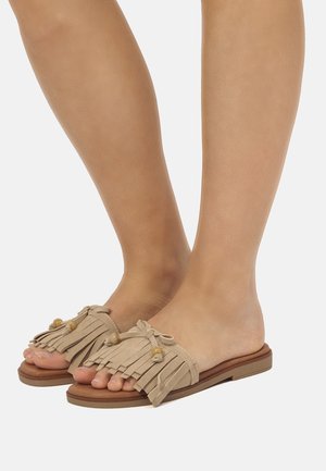 Pies con sandalias beige tipo slide con flecos y adornos de cuentas sobre suelas planas marrones, sobre un fondo blanco.