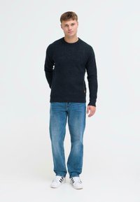 Tume navy sinise kaeluse jumper, tekstureeritud kangast, koos helesiniste sirgete pükstega ja valgete tossudega mustade triipudega.