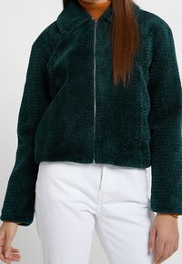 Grøn fleecejakke, cropped design, med krave og lynlås lukning. Blød tekstur, lange ærmer, parret med hvide bukser.