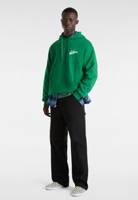 Felpa verde con cappuccio e tasca frontale, abbinata a una maglietta grigia, pantaloni neri e scarpe slip-on a scacchi. Sfondo semplice.