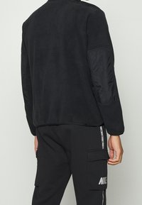 Schwarze Jacke mit einer weichen Textur, ausgestattet mit dunkelblauen Polyesterärmeln, einem hohen Kragen und einem lässigen, cropped Design.