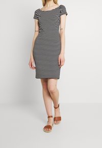 Robe rayée noir et blanc à manches courtes et décolleté carré, fabriquée dans un tissu léger. Portée avec des sandales marron à lanières.