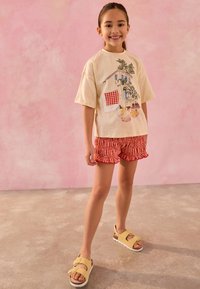 T-shirt ivoire avec un motif coloré de maison et de fruits, accompagné d'un short à carreaux rouges. Porte des sandales jaunes avec des semelles blanches.