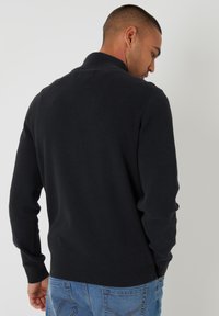 Czarny dziany sweter z wysokim kołnierzem i prążkowanymi mankietami. Ponadczasowy fason o gładkiej fakturze, noszony z niebieskimi dżinsami.