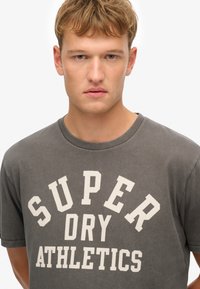 Camiseta de algodón gris con un gran gráfico blanco que dice "SUPER DRY ATHLETICS" en una tipografía serif en negrita. Cuello redondo clásico y mangas cortas.