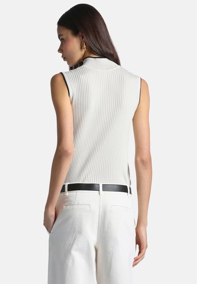 Femme aux longs cheveux foncés portant un haut blanc sans manches côtelé et un pantalon blanc avec ceinture noire, vue de dos.