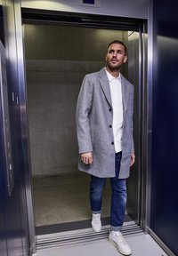 Cappotto di lana grigio con bottoni, camicia bianca, jeans blu e sneakers bianche, in piedi in un ascensore con uno sfondo di cemento.