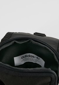 Borsa nera con zip dal texture liscio e opaco. All'interno presenta una fodera verde e un'etichetta bianca con il logo Adidas e il testo.