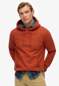 Rust-colored hoodie with a kangaroo pocket, drawstring hood, and embroidered logo; layered over a blue and green plaid shirt.