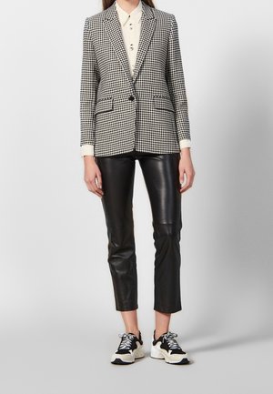 Osoba w czarno-białej marynarce w kratę typu pepitka, białej koszuli na guziki, czarnych skórzanych spodniach o długości 7/8 oraz czarno-białych trampkach, stojąca.