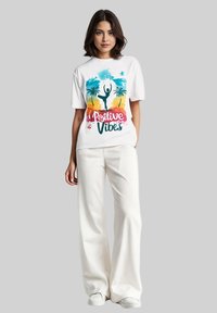 Witte katoenen t-shirt met een kleurrafische afbeelding van palmbomen en een gesilhouetteerde figuur. Gecombineerd met hoge witte wijde broek en sneakers.