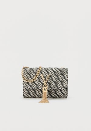 Bolso de mano tejido en blanco y negro con cadena dorada, cierre en forma de V dorado y detalle de borla sobre un fondo liso.