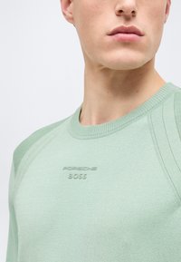 Κοντινό πλάνο ενός άνδρα που φοράει ένα ανοιχτοπράσινο πουλόβερ με στρογγυλή λαιμόκοψη, με την επιγραφή "Porsche BOSS" ανάγλυφη στο στήθος.