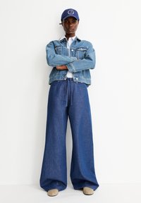 Hellblaujeansjacke mit zwei Taschen, kombiniert mit weit geschnittenen dunkelblauen Jeans und hellbraunen Schuhen. Marineblaue Mütze mit Logo auf der Vorderseite.