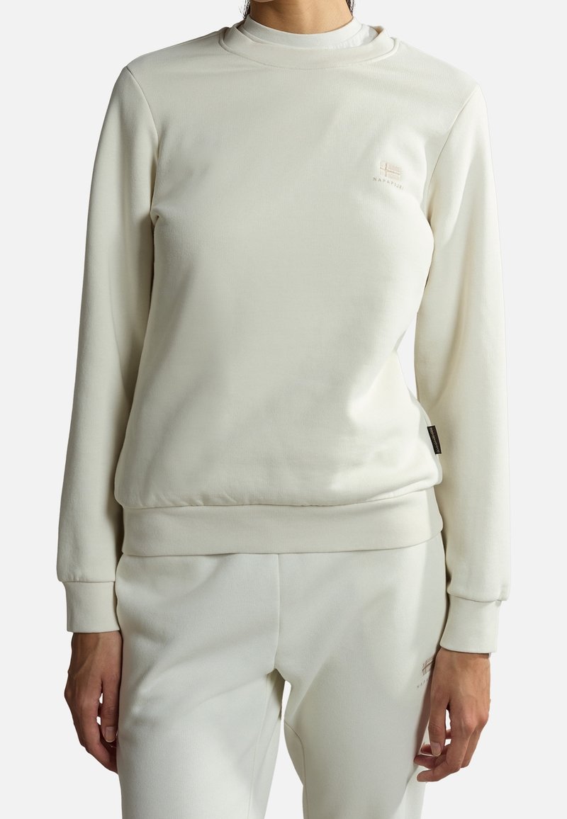 Femme portant un sweat-shirt à manches longues couleur écru et un pantalon assorti, debout les bras détendus le long du corps sur un fond blanc.