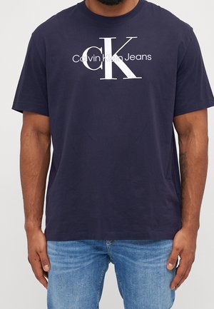 Mann trägt ein marineblaues Calvin Klein Jeans T-Shirt mit großem weißem "cK" Logo und Text, kombiniert mit hellblauen Jeans.
