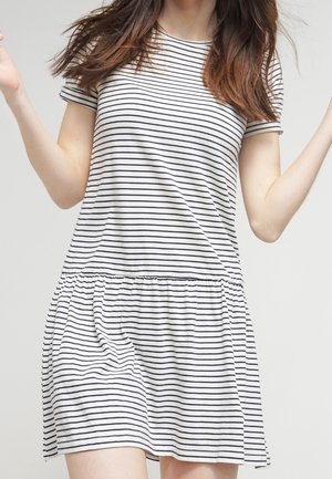 Femme portant une robe blanche à manches courtes avec de fines rayures horizontales noires, debout devant un fond clair uni.