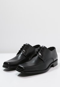 Chaussures habillées en cuir noir avec une finition lisse et des détails perforés sur les côtés. Caractérisées par un bout pointu et un design à lacets.